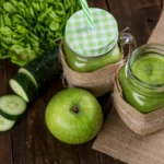 Copo de vidro com suco verde vibrante, decorado com uma fatia de maçã verde e folhas de hortelã sobre mesa clara.