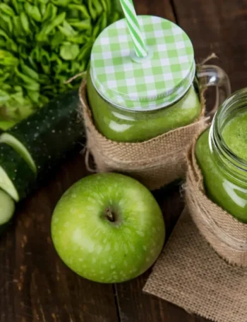 Copo de vidro com suco verde vibrante, decorado com uma fatia de maçã verde e folhas de hortelã sobre mesa clara.
