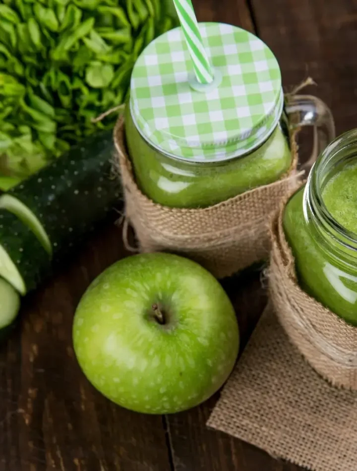 Copo de vidro com suco verde vibrante, decorado com uma fatia de maçã verde e folhas de hortelã sobre mesa clara.
