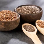 Duas tigelas pequenas de madeira, uma com sementes de chia pretas e outra com linhaça dourada, sobre uma mesa clara.