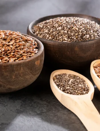 Duas tigelas pequenas de madeira, uma com sementes de chia pretas e outra com linhaça dourada, sobre uma mesa clara.