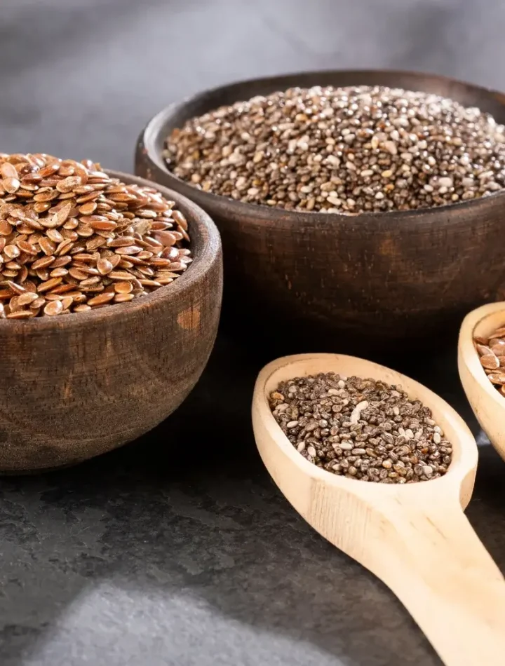 Duas tigelas pequenas de madeira, uma com sementes de chia pretas e outra com linhaça dourada, sobre uma mesa clara.