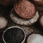 Grãos de cores variadas dispostos em colheres de madeira sobre fundo neutro.