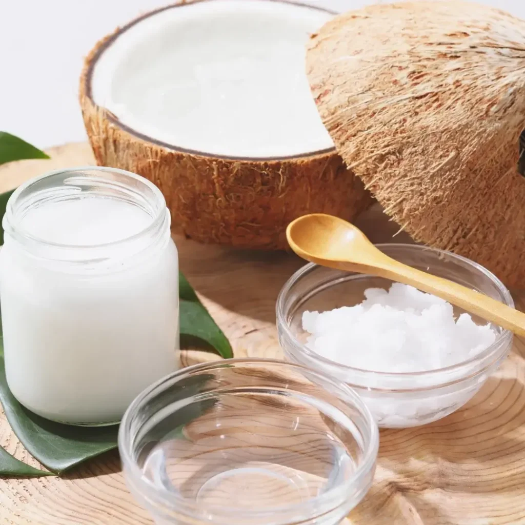Pote de vidro com óleo de coco sólido branco e pedaços de coco seco ao redor.