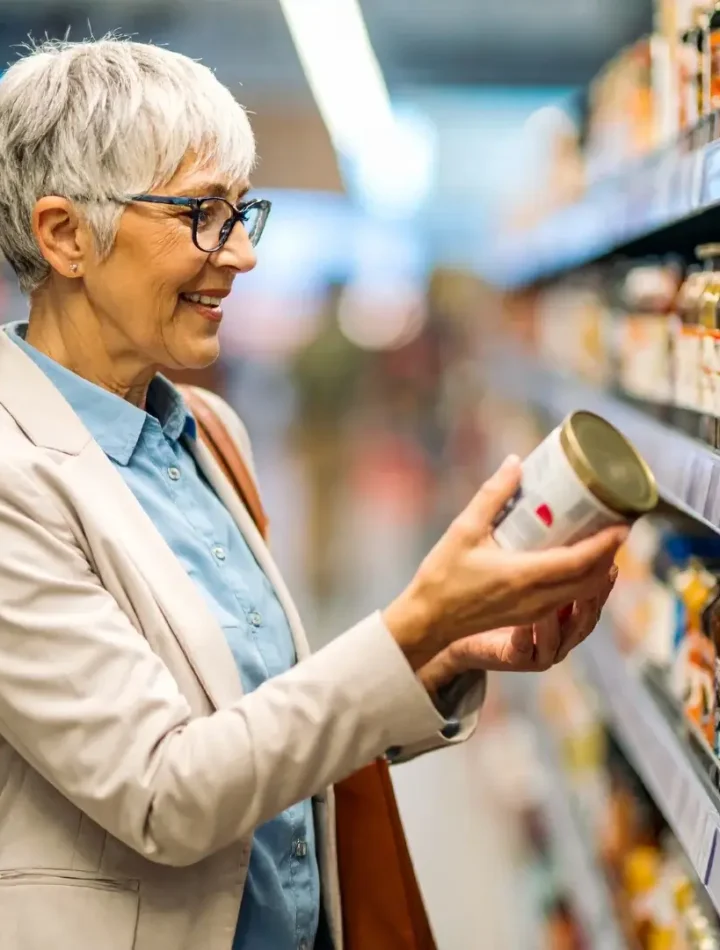 Uma pessoa segurando uma embalagem de alimento e lendo atentamente as letras pequenas no verso em um corredor de supermercado.