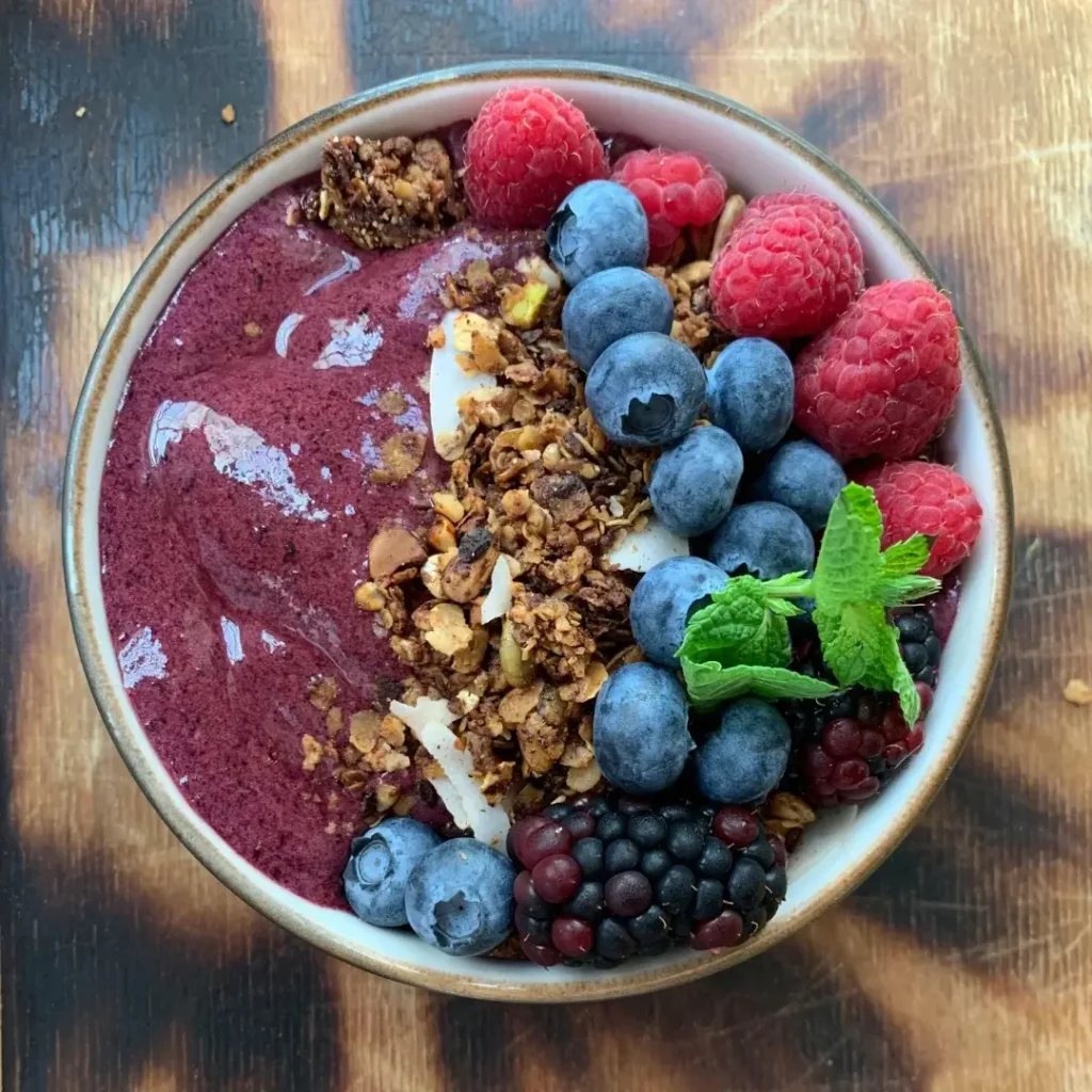 Uma tigela de café da manhã com creme roxo, decorada com fileiras de mirtilos, fatias de morango e sementes de chia.